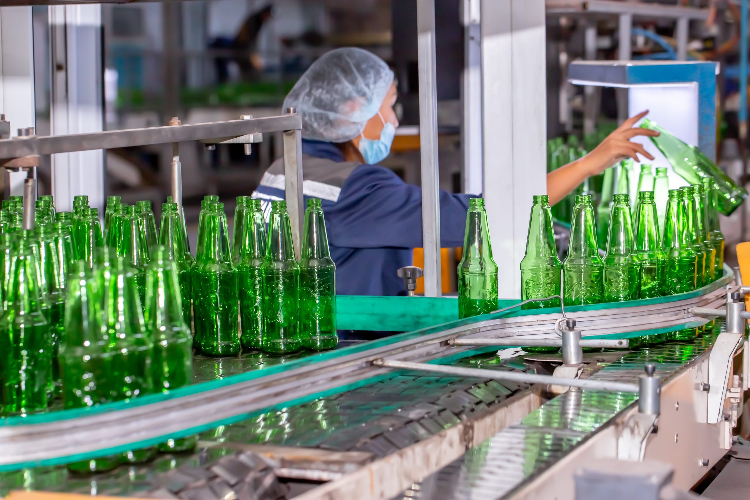Glass manufacturing plant worker monitoring production line with green glass bottles, bulk material handling system in operation